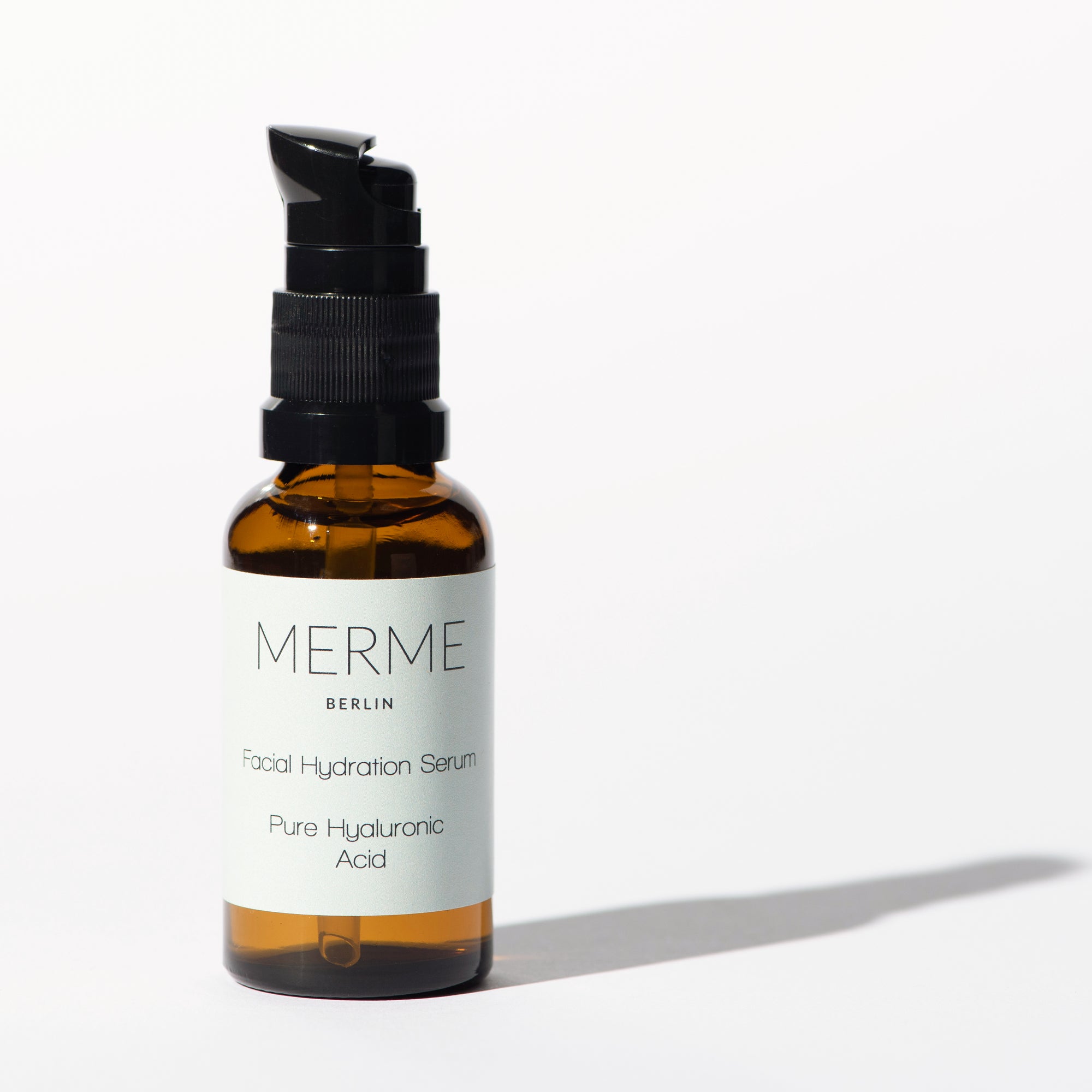 A brown glass bottle with a black pump dispenser labeled Merme Berlin Hyaluron Serum is displayed on a white background, emphasizing the brand Merme and its benefits for intense hydration and moisture retention.