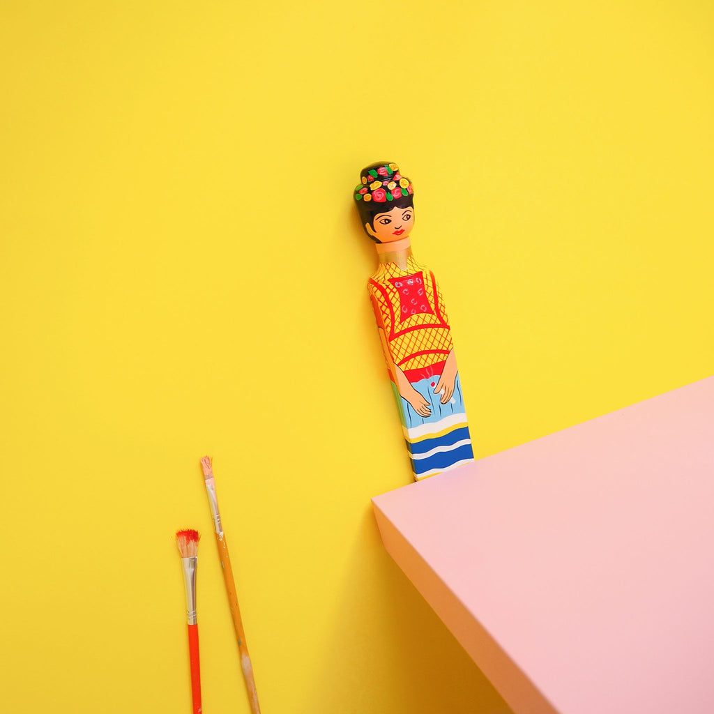 The Wedgie FRIDA Door Stopper by Wedgie, a hand-painted Alstonia wood piece inspired by Frida Kahlo, rests on a yellow background next to two paintbrushes; a pink rectangular surface appears in the lower right corner.