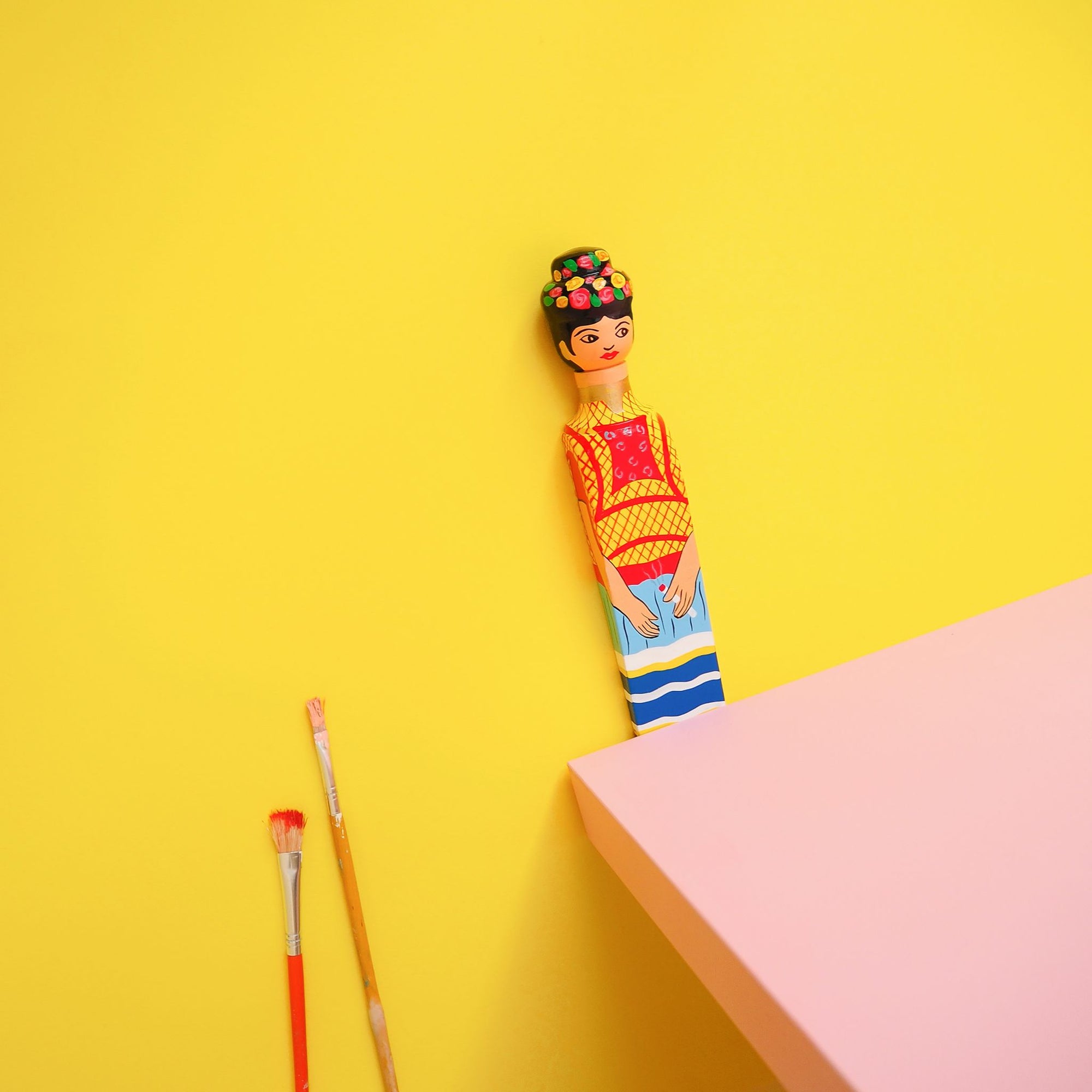 The Wedgie FRIDA Door Stopper by Wedgie, a hand-painted Alstonia wood piece inspired by Frida Kahlo, rests on a yellow background next to two paintbrushes; a pink rectangular surface appears in the lower right corner.